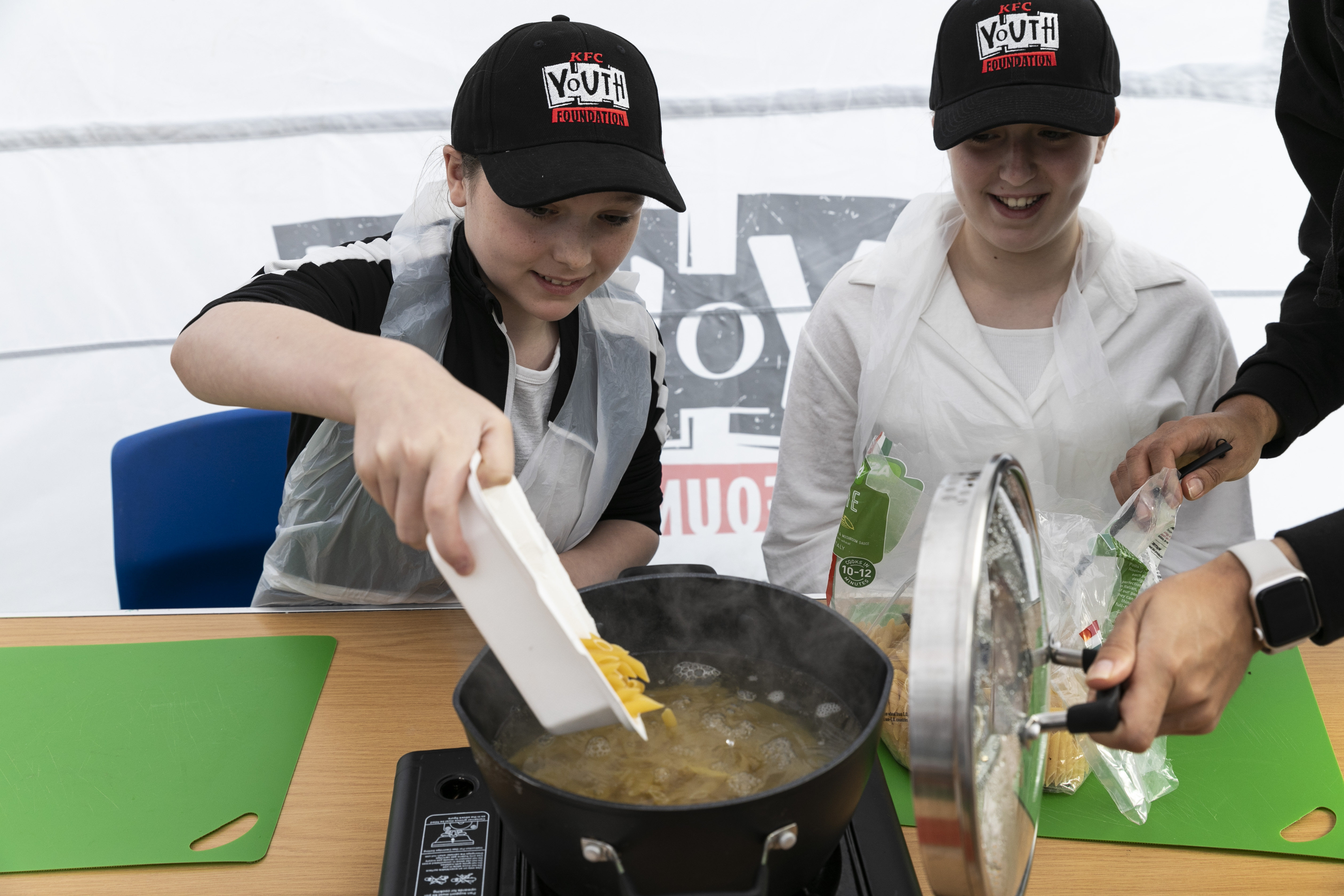 One girl pouring dry pasta into a pot with hot water, another girl watching on, both smiling. Another person off camera holding lid of pot. KFC Youth Foundation banner in the background